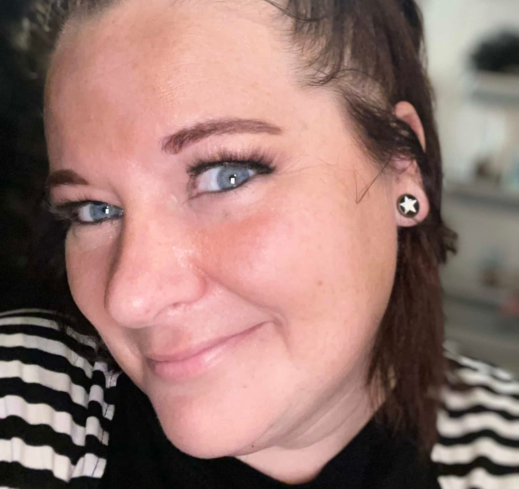 Close-up portrait of a smiling person with blue eyes, star earrings, and a black-and-white striped top