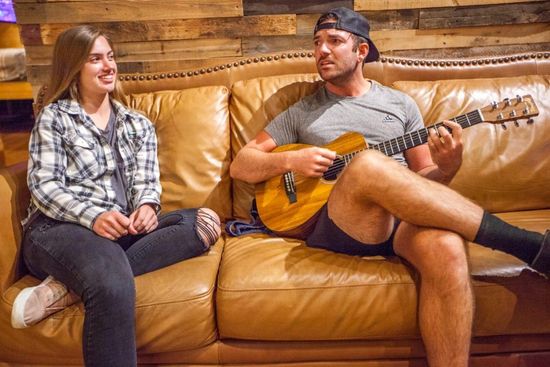 Two people on a tan couch, smiling; one holds an acoustic guitar in a cozy living room.