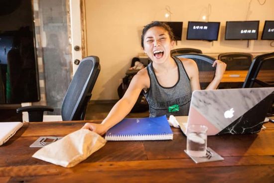 Person laughing at a wooden table with a laptop, notebook, drink, and paper bag in a gym-like room.