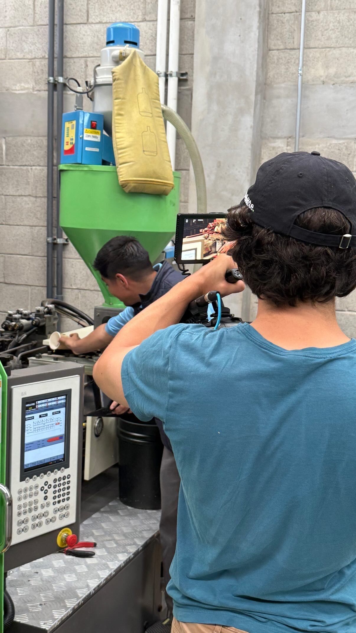 Person in blue shirt photographing a worker in a metal workshop beside a machine and tools
