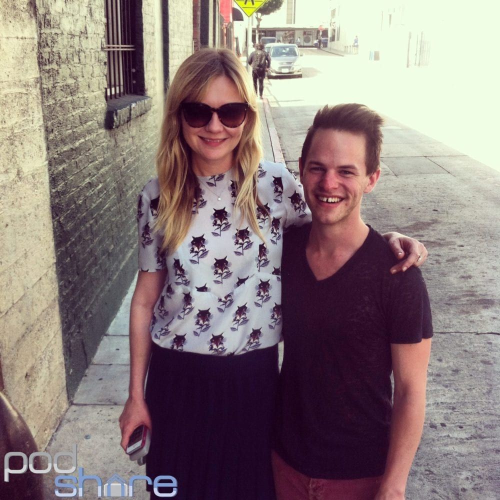 Actress Kirsten Dunst with sunglasses and a man smile and pose for a picture, arm in arm, on a sidewalk.