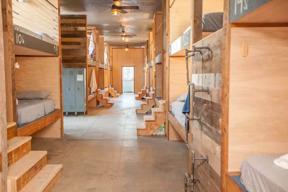Dorm room with bunk beds, wood accents, and blue lockers. Sunlight streams down a central hallway.