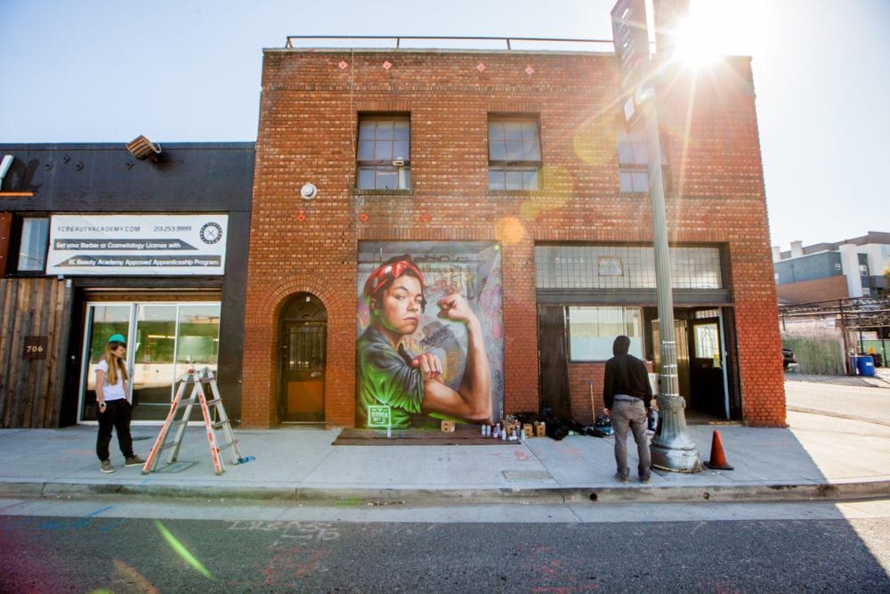 Street mural of a woman flexing her arm. Artists work on the building. Bright sun.