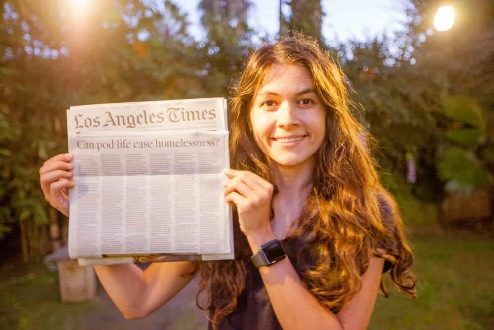 Woman smiling outdoors, holding up a Los Angeles Times newspaper at dusk