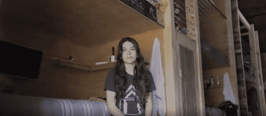 A young woman stands in a small room with bunk beds, looking at the camera.