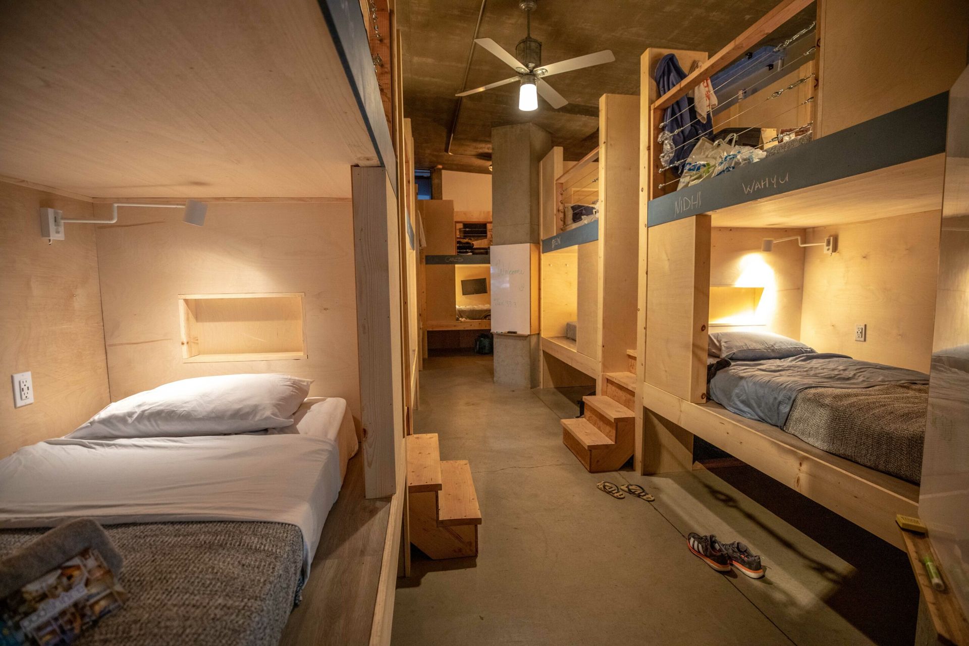Dorm room with bunk beds and single beds. Wooden structures, concrete walls, and a ceiling fan.
