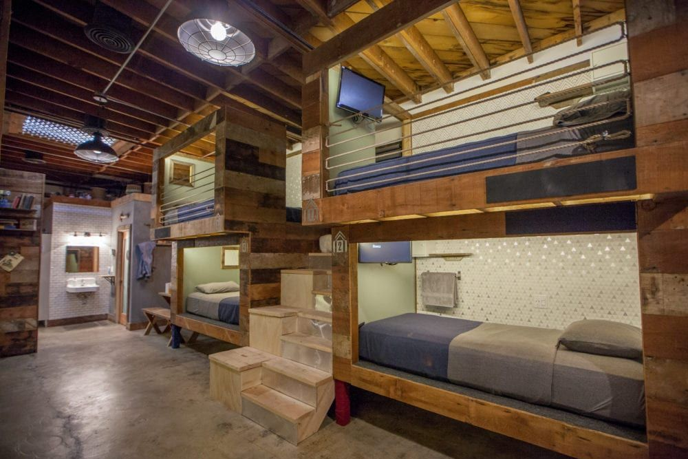 Loft-style dorm room with bunk beds, wood beams, and a staircase in an industrial interior