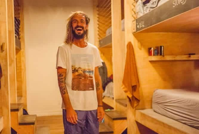 Man standing in a warm-lit bunk room with wooden bunks, bedsheets, and a towel on the wall.