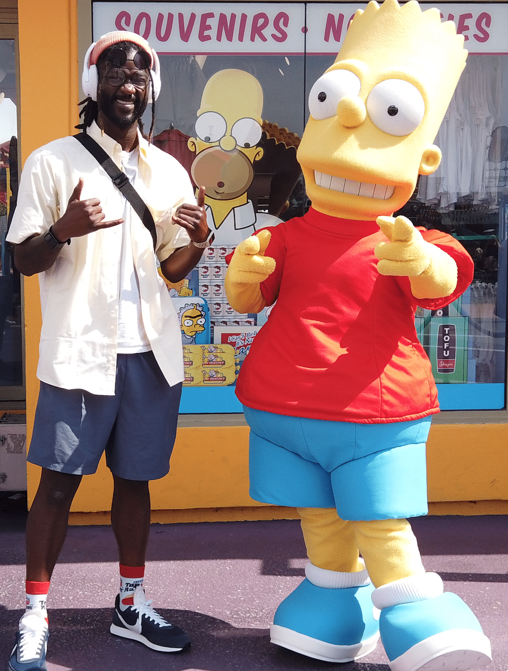 Man posing beside a Bart Simpson mascot outside a souvenir shop.