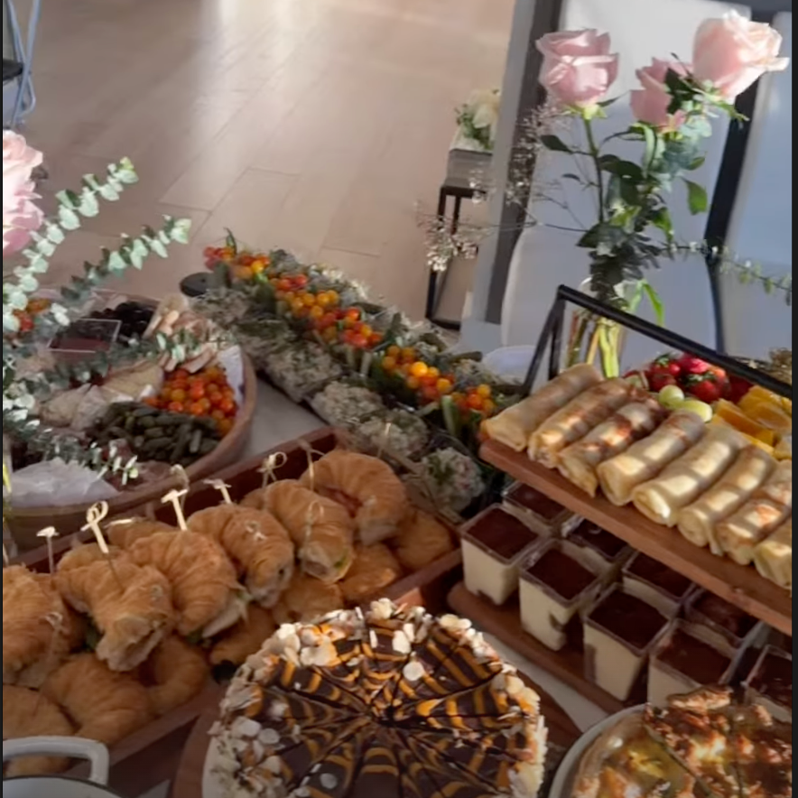 Buffet table with pastries, fruit, and desserts arranged indoors near pink flowers