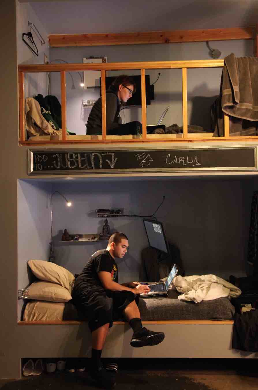 Two people in a small space. One on a loft, the other on a bed, both on laptops.
