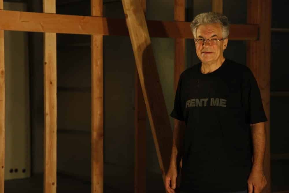 Man in black shirt stands in front of a wooden wall frame. 
