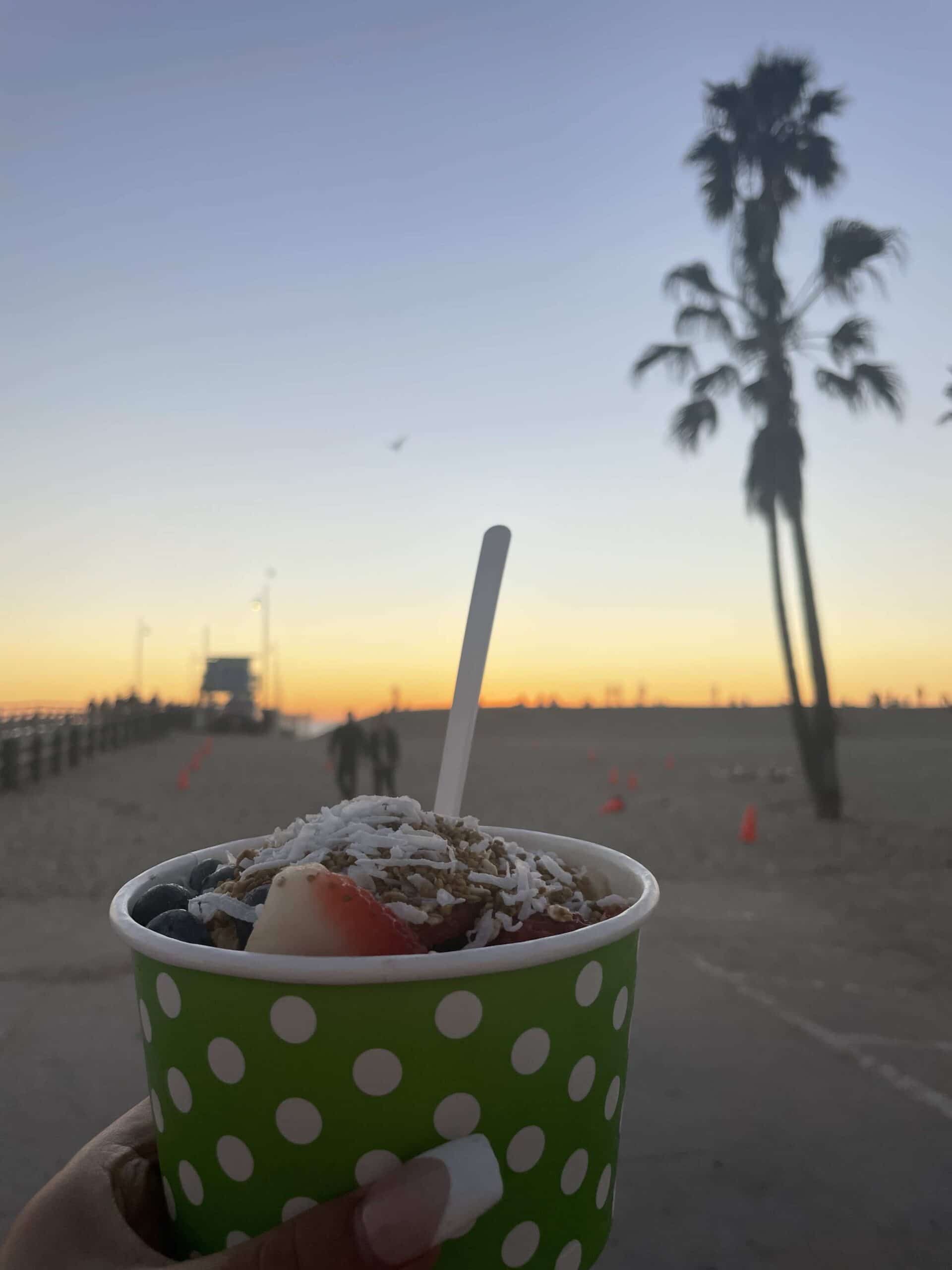 a hand holding a green polka-dot cup filled with a dessert topped with fruit and granola, with a beach and palm tree in the background at sunset
