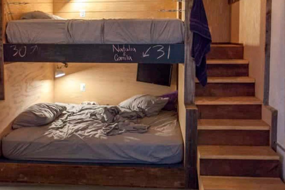 A bunk bed with a lower mattress and staircase, in a warm wooden room with handwritten labels on the frame.