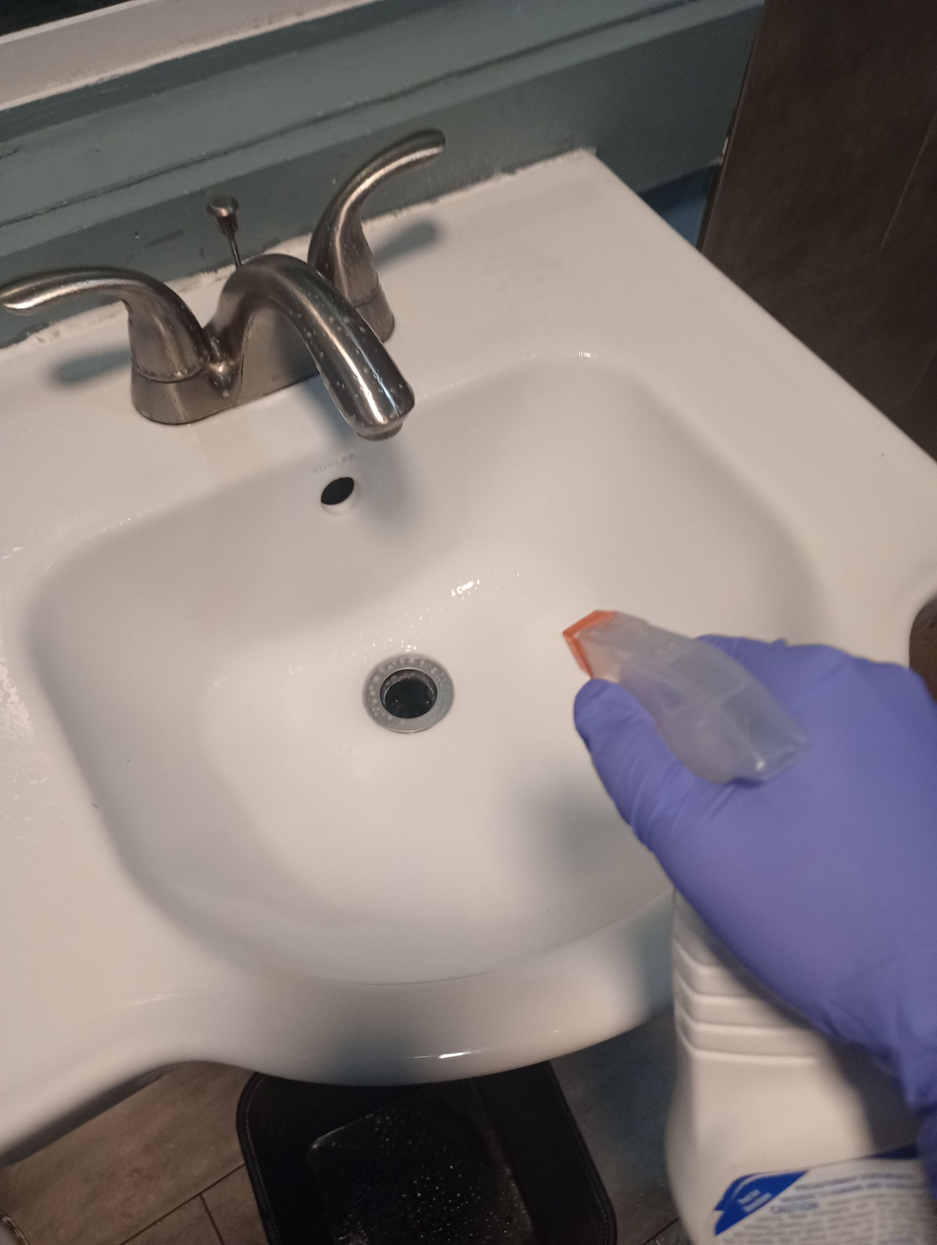 Gloved hand spraying cleaner into a white bathroom sink with chrome faucet