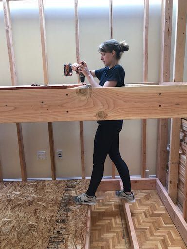 Woman using a drill on wood beam in a room under construction. She wears black leggings, grey shoes.
