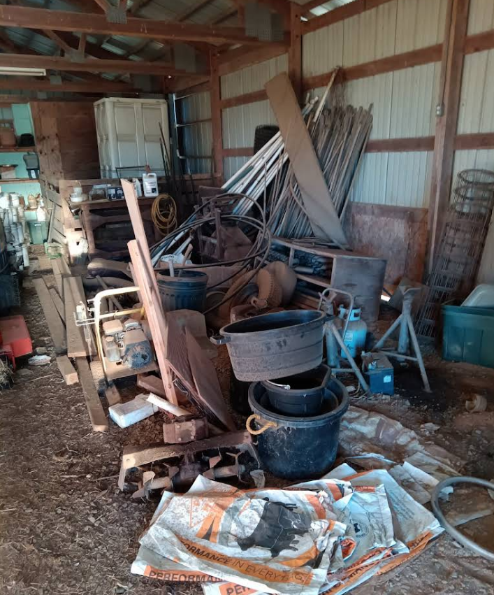 Cluttered shed interior with piles of scrap metal, tools, buckets, wood, and various debris.
