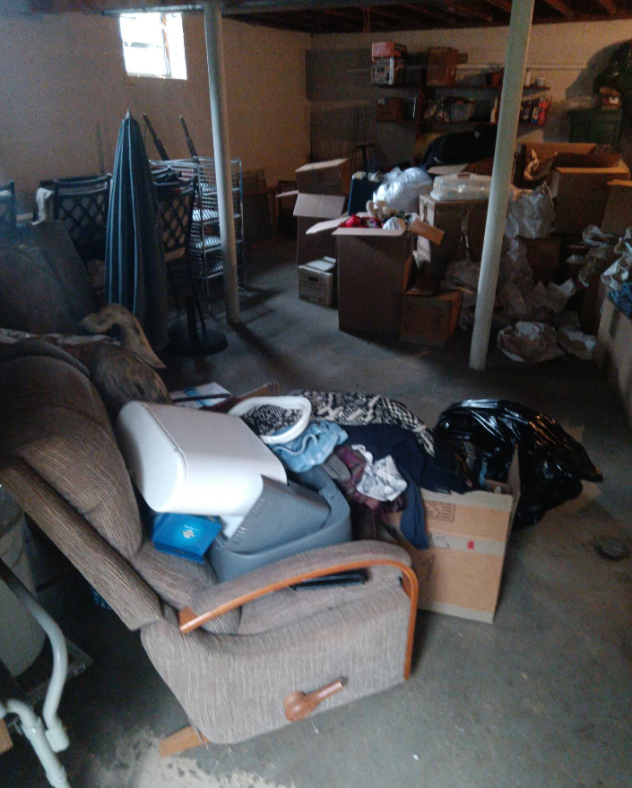 Cluttered basement with a recliner chair, boxes, and various items scattered around.