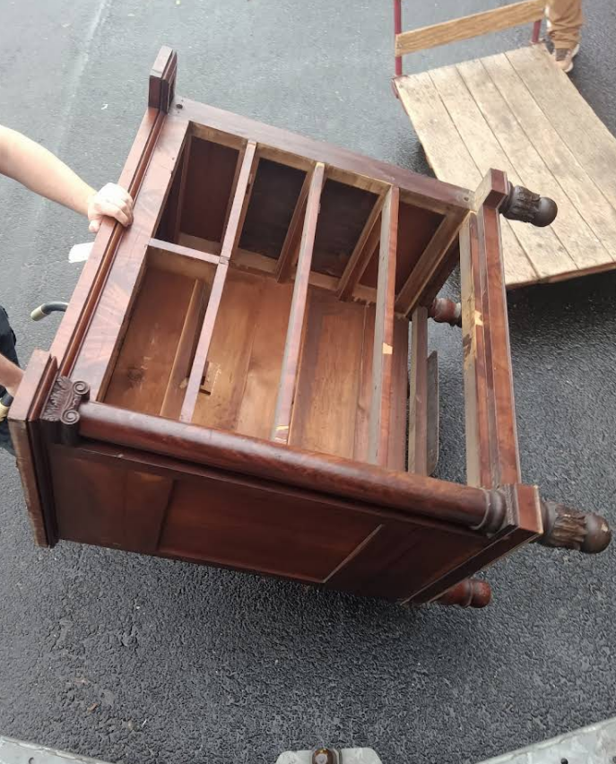 A dark wood, empty antique nightstand being moved on pavement; person's hand visible.