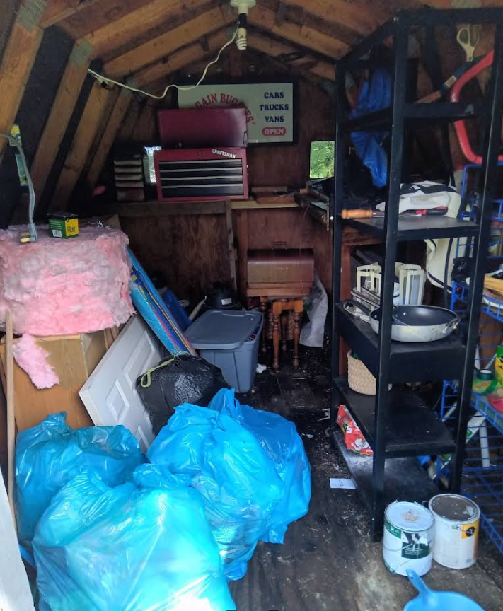Interior of a cluttered shed with tools, shelving, and bags of trash. Dark wood, with blue and pink items.