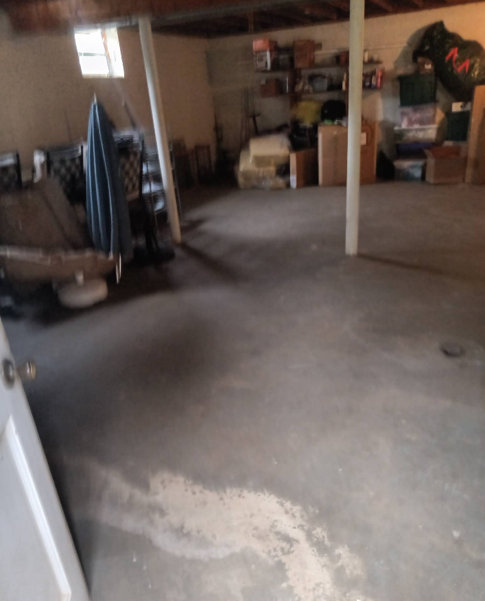 Basement with concrete floor and clutter. White door in foreground. Window in background.