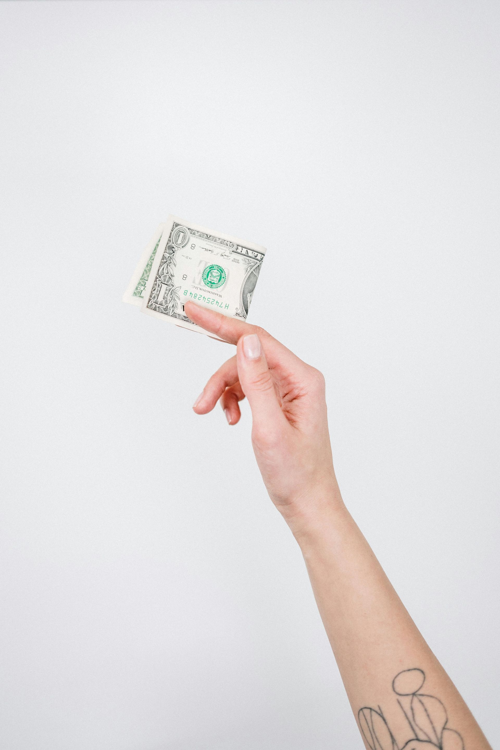 Hand holding a single dollar bill against a white background. Tattoo on forearm.