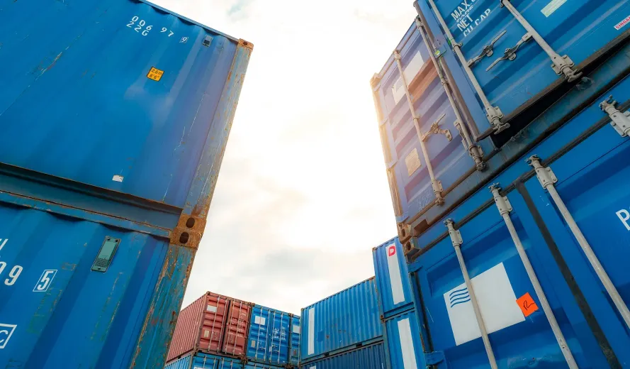 Stacked blue shipping containers at a port, viewed from below against the sky