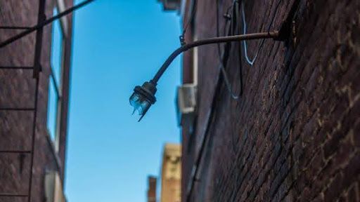 A broken, dangling outdoor light fixture attached to a brick wall in a narrow urban alleyway under a bright blue sky.