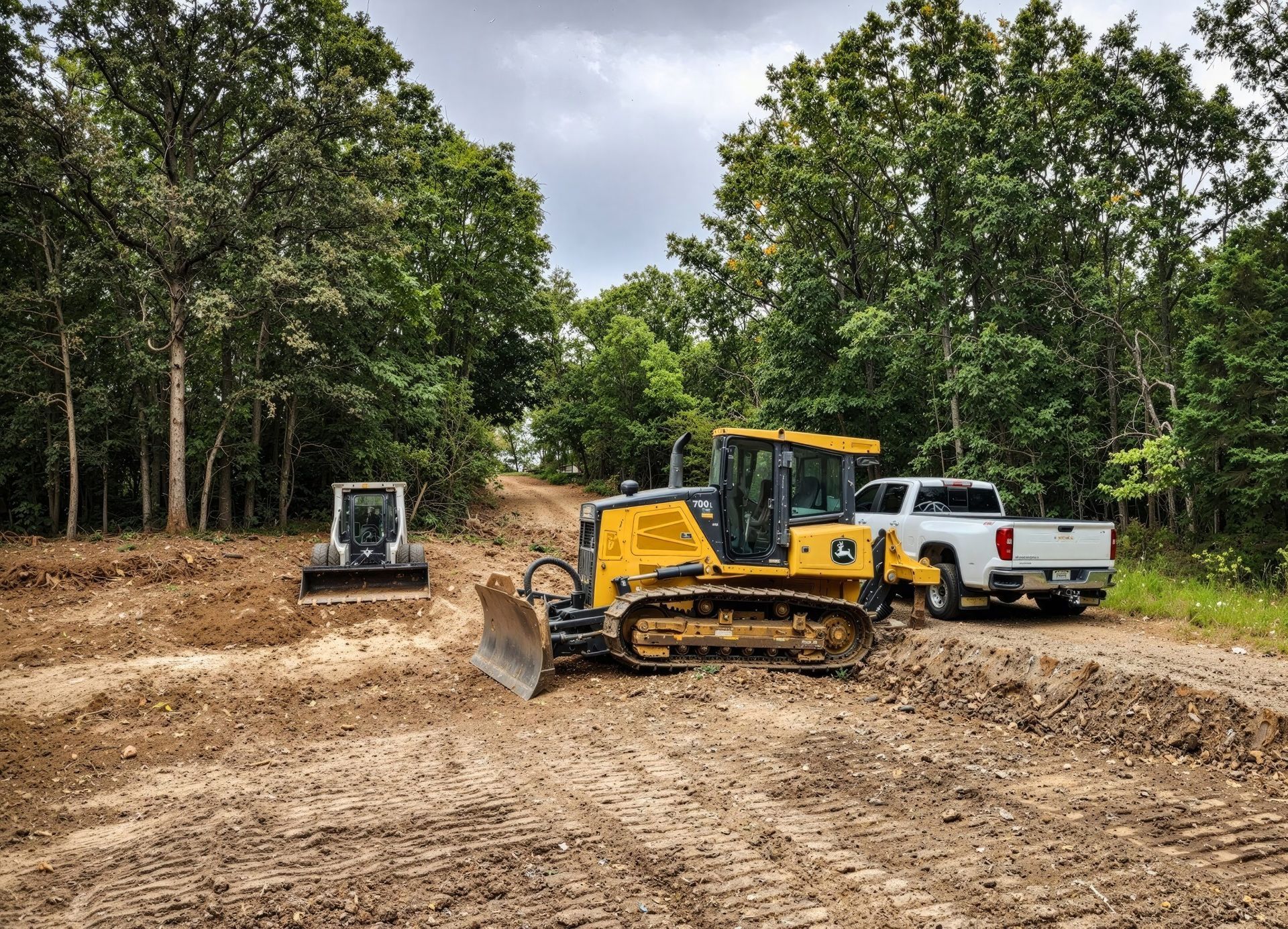 Bulldozer grading a muddy road beside a white pickup and another machine in a wooded area