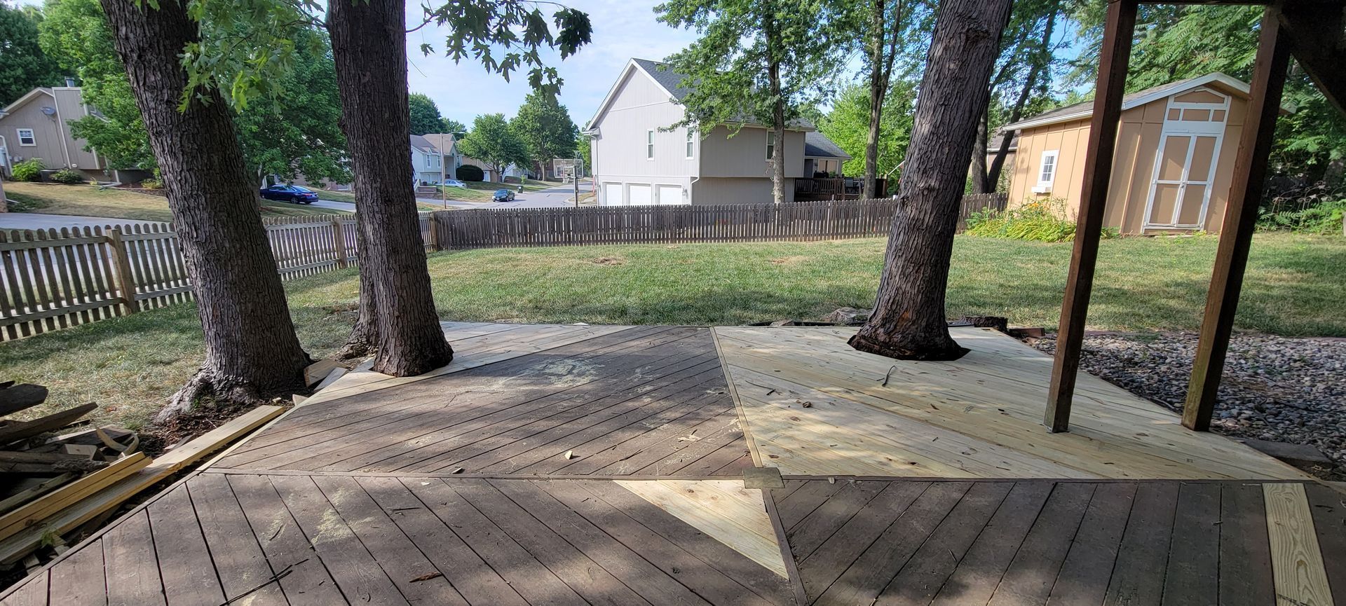 A backyard deck with two trees growing through it. A small shed and houses are in the background.