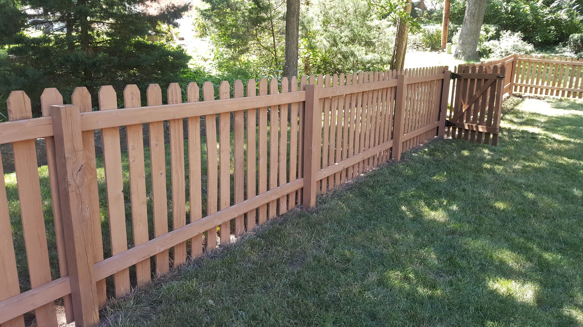Brown picket fence in a grassy yard, with trees in the background.