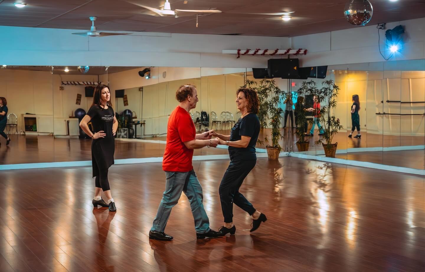 People dancing in a studio; a woman watches them. Bright dance floor, mirrors, and decorations.