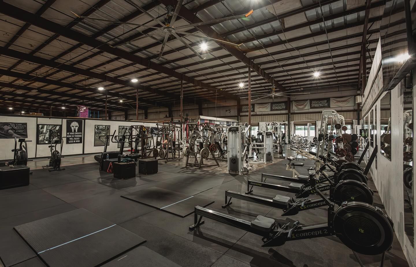 Large, open gym with various workout equipment, including rowers and weight machines. Metal roof, black flooring.
