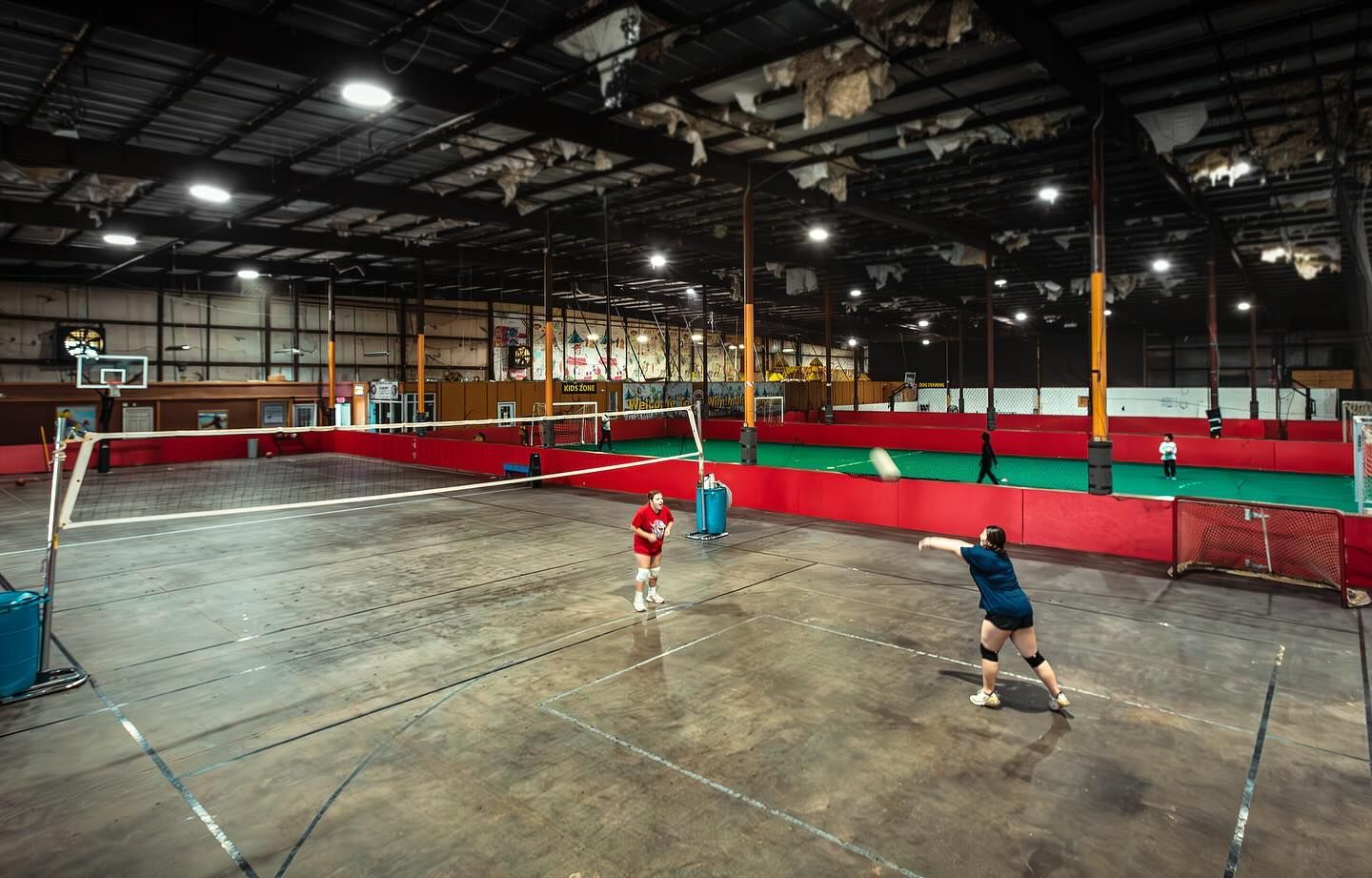Indoor volleyball game in progress; players in red and blue uniforms, concrete floor, red and green walls.