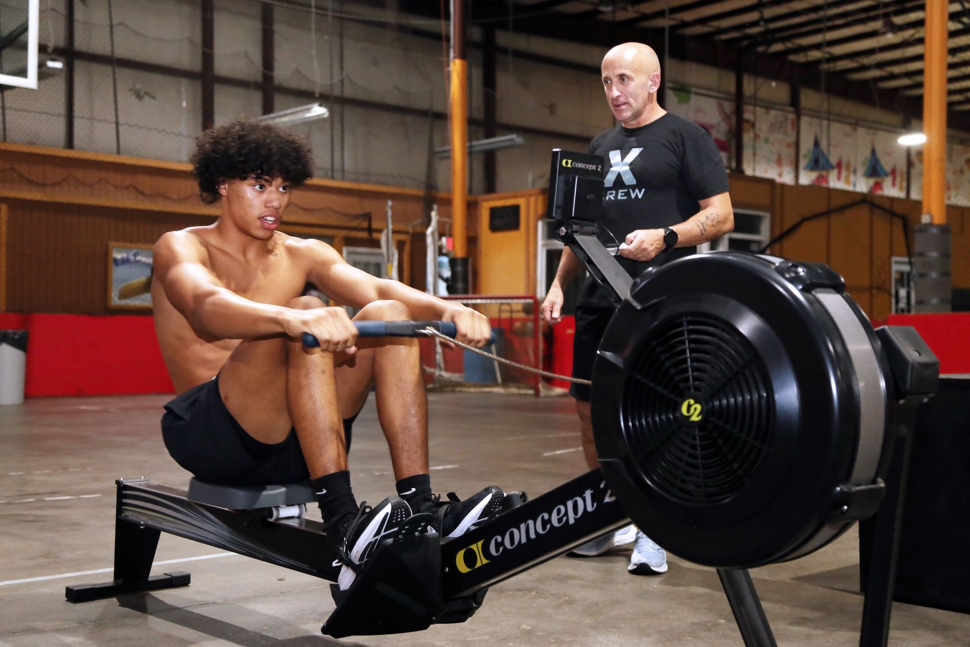 Young man rows on a Concept 2 machine, supervised by a coach in a gym setting.