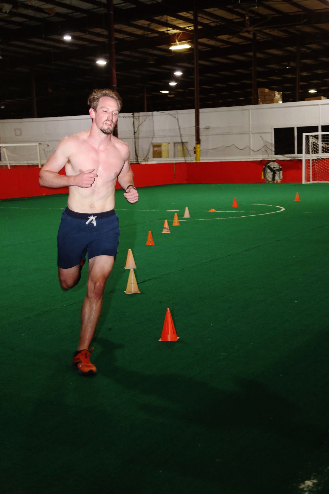 Man running toward the camera, passing orange cones on a green indoor field. He is shirtless with blue shorts.