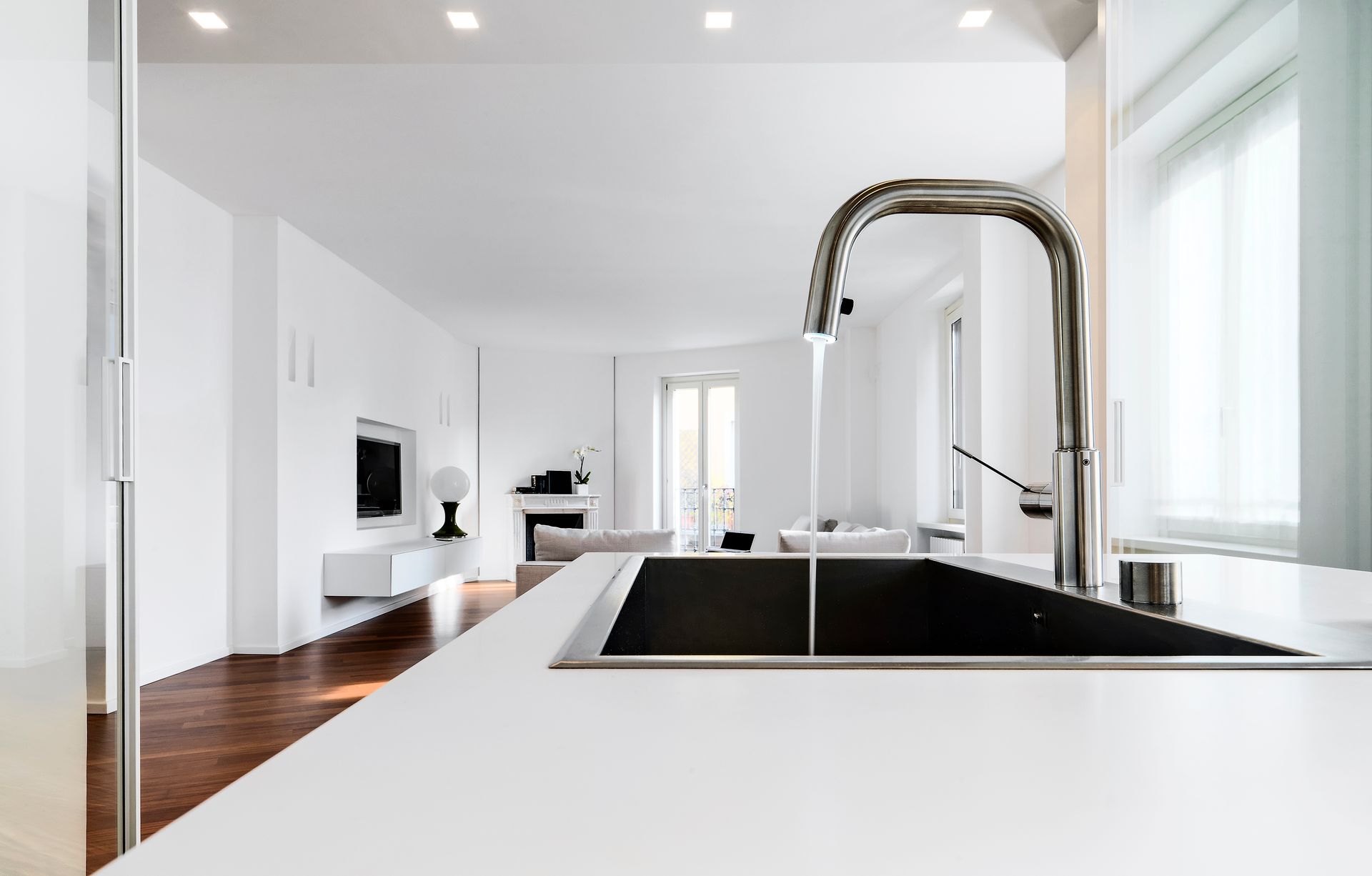 Robinet chromé avec eau courante au-dessus d'un évier noir dans une cuisine moderne blanche, menant au salon.