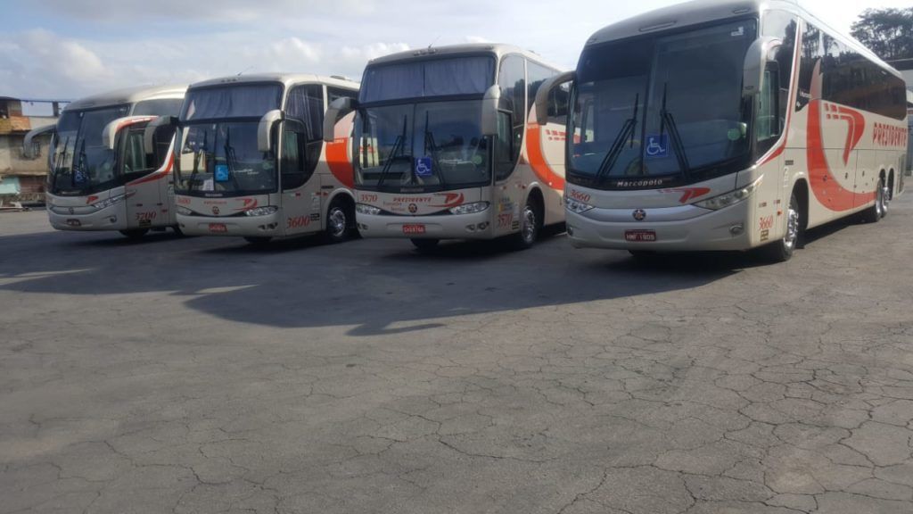 Quatro ônibus turísticos brancos e laranjas estacionados lado a lado em um terreno pavimentado.