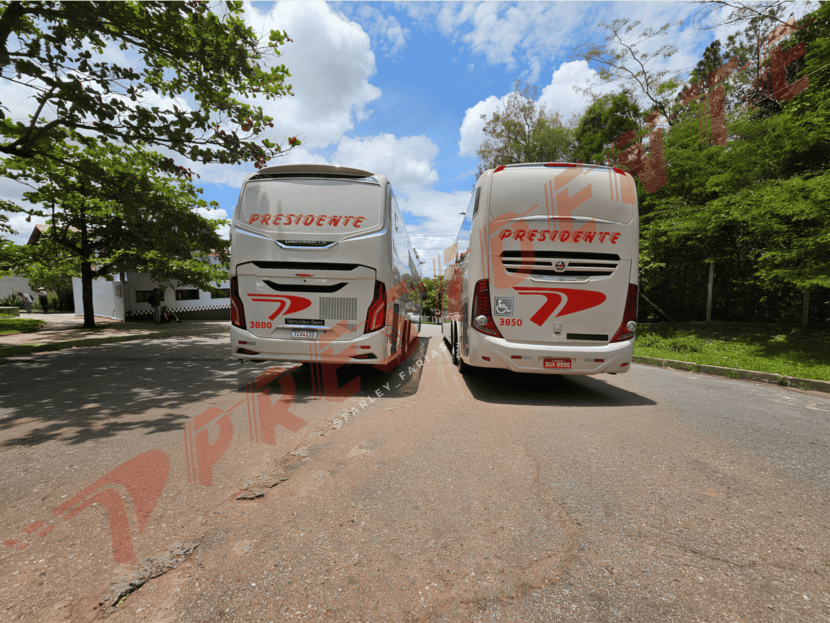 Duas autocaravanas brancas com detalhes em vermelho estacionadas lado a lado numa estrada arborizada sob um céu azul.