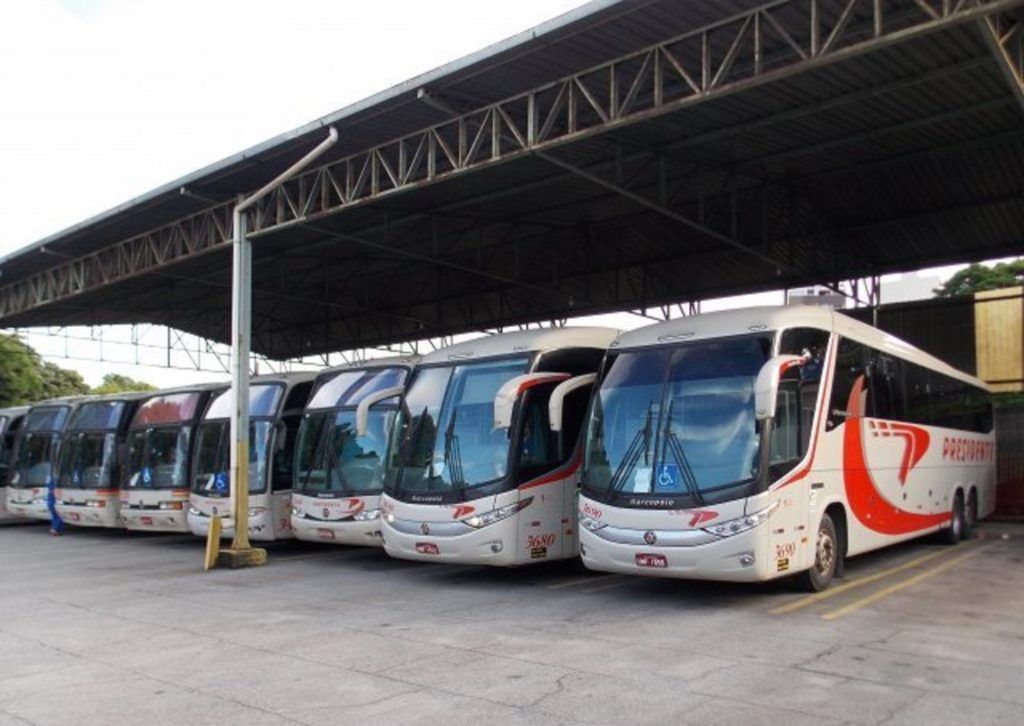Fileira de ônibus turísticos brancos e vermelhos estacionados sob um grande depósito coberto.
