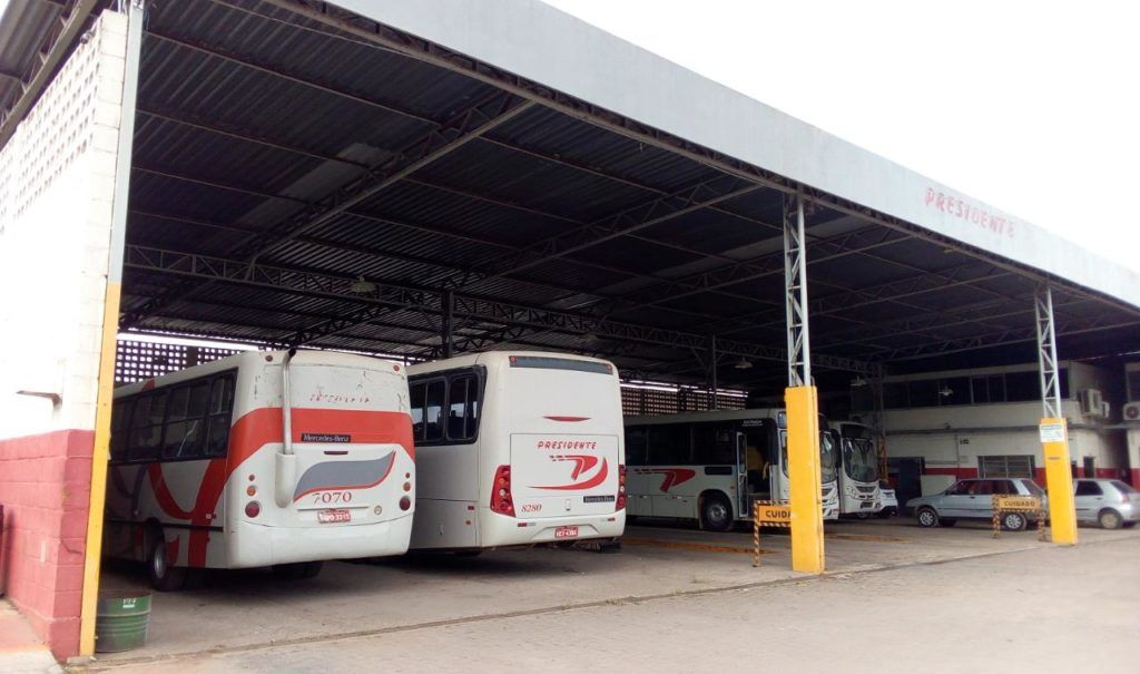 Área coberta de estacionamento para ônibus com vários ônibus brancos e amarelos estacionados em uma garagem.