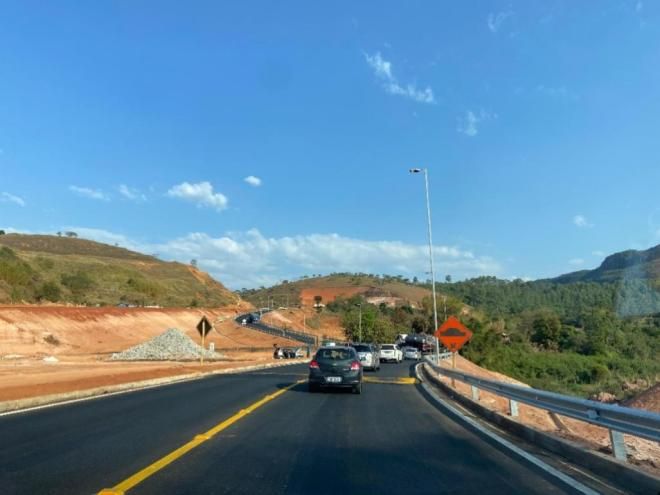 Carros trafegando em uma rodovia sinuosa através de uma área de construção montanhosa sob um céu azul claro.