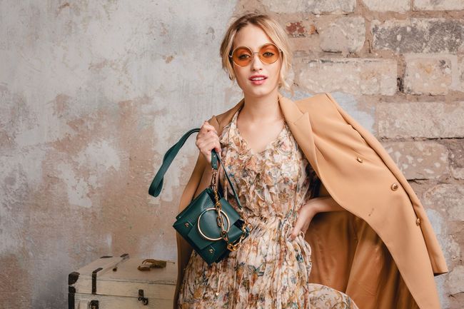 Woman in floral dress and tan coat, holding a teal handbag against a textured wall.