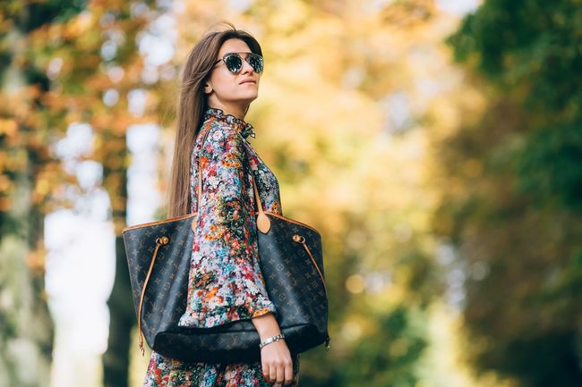 Woman in sunglasses carrying a large tote bag, standing on a sunlit tree-lined path.