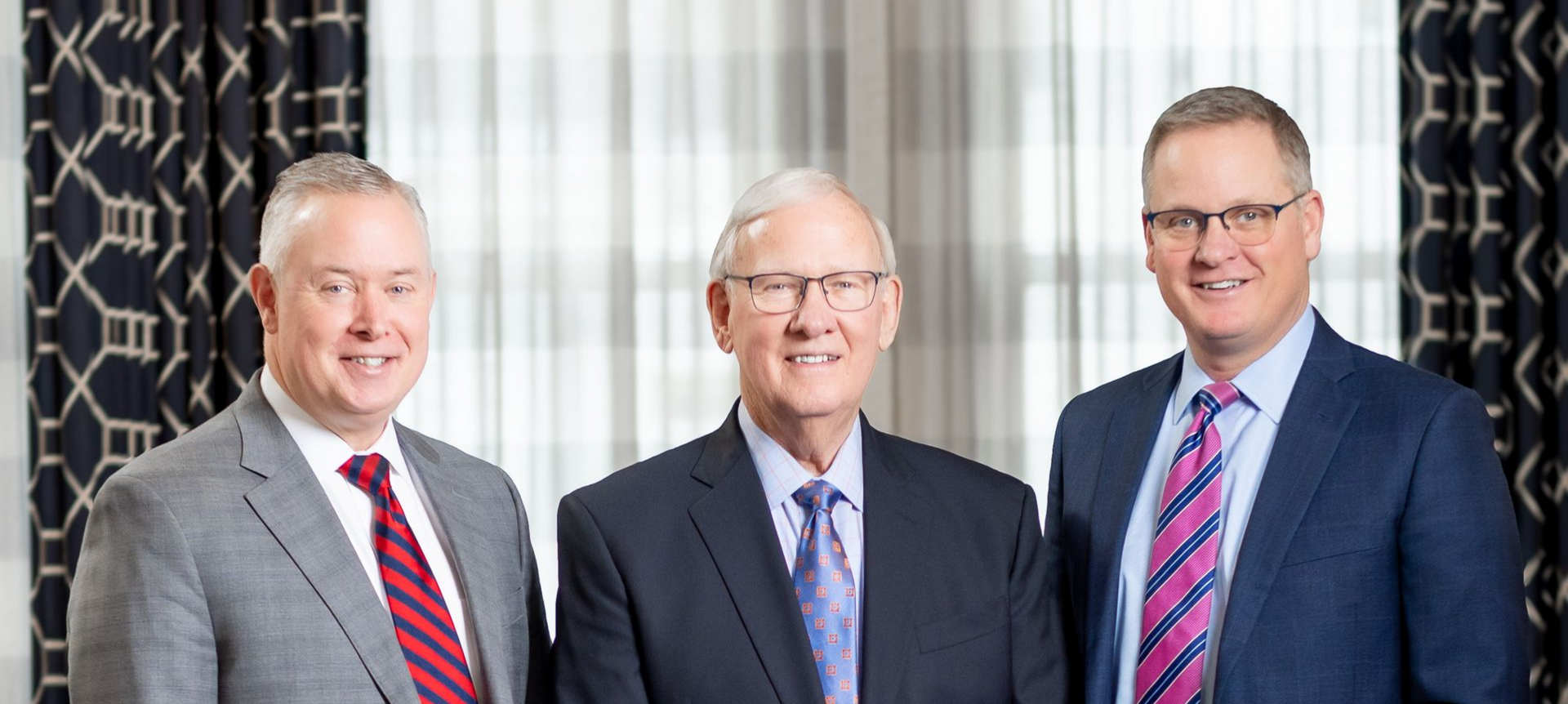 Three men in suits standing together indoors in front of striped curtains.
