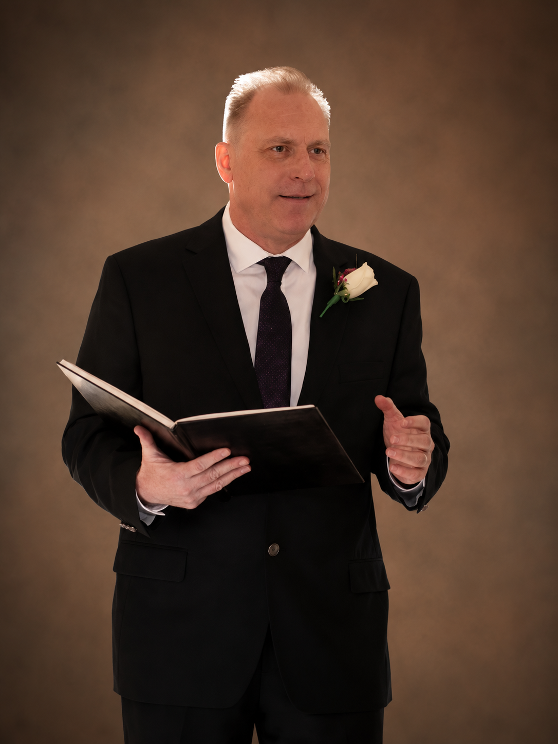Man in a black suit holding an open book, speaking against a warm studio backdrop.