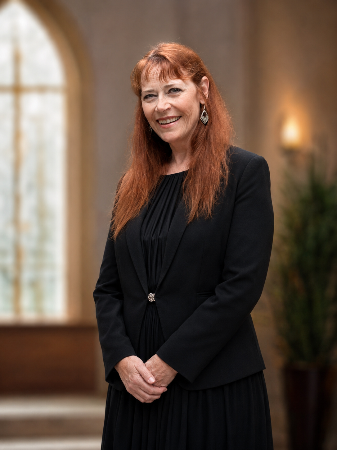 Woman in a black dress and blazer, smiling in a softly lit room with an arched window.