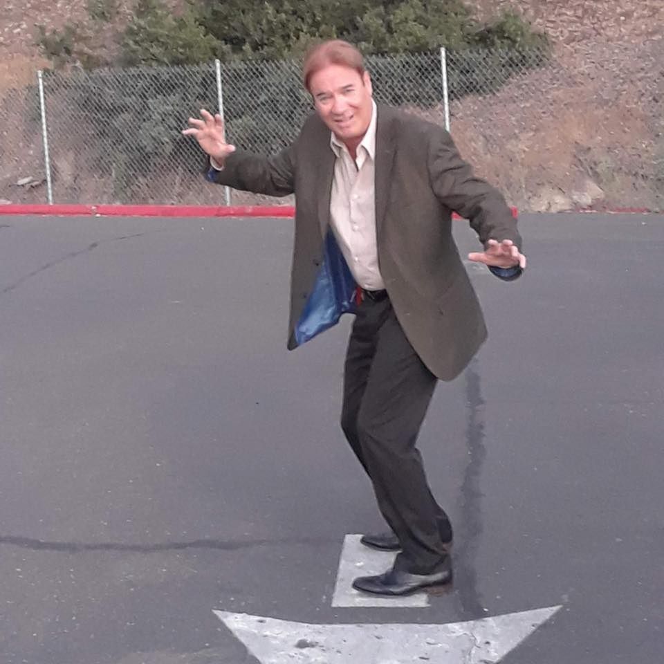 Man posing on a painted arrow in a parking lot, arms out, near a chain-link fence and hillside.