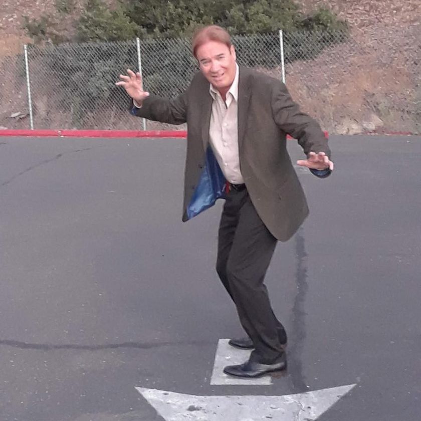 Man balancing on a skateboard in a parking lot, arms out, wearing a blazer and dress shoes