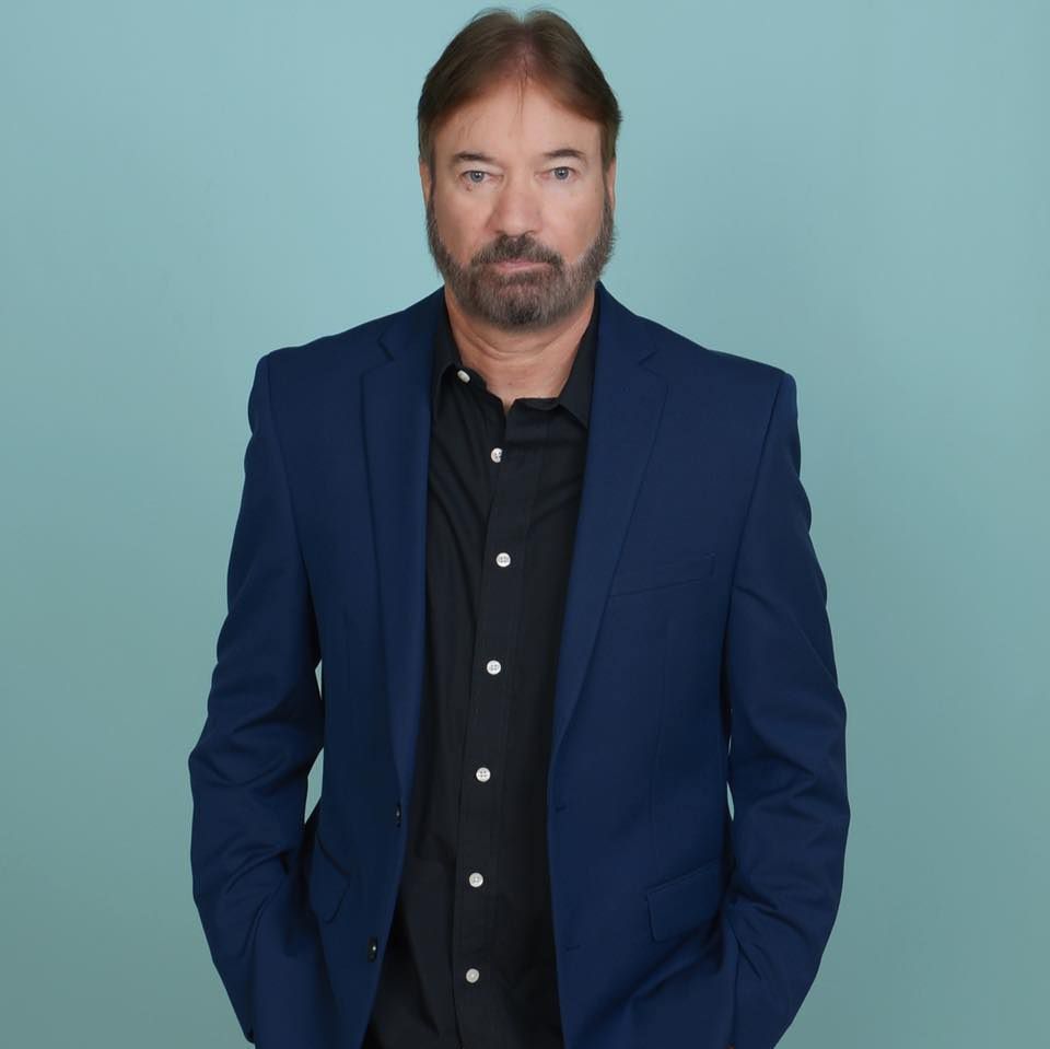 Man in a navy blazer and black shirt standing against a light blue background.